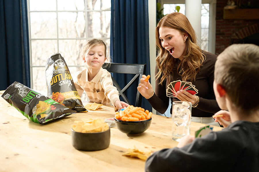 Family sharing Outlaw Snax Spicy Ranch tortilla chips and Space Puffs cheese snacks while playing cards.